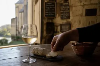 Subtle detail of a hand arranging cheese next to a glass of Saumur blanc on an old wooden table, golden afternoon light streaming into a rustic tasting room with a blurred stone cellar in the background.