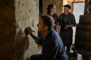 Three generations of a winemaking family in a historic tuffeau wine cave, with one member revealing an old engraving and two others in soft focus, bathed in natural morning light.