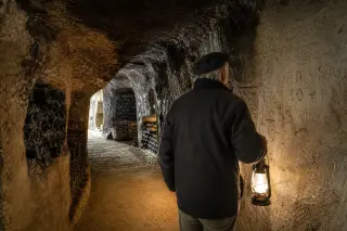Elderly vintner in a tuffeau cave studying ancient wall inscriptions by lantern light, wine bottles resting in stone niches with soft golden illumination.